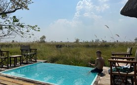 Ein Junge sitzt am Rand eines Pools und spielt mit Wasser, umgeben von hohen Gräsern und einem klaren Himmel.