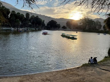 Am Ufer eines ruhigen Sees sitzen zwei Personen und genießen den Sonnenuntergang hinter den Sanddünen.
