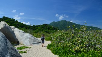 Ein Wanderer geht barfuß auf einem sandigen Pfad, umgeben von großen Felsen und üppiger grüner Vegetation.