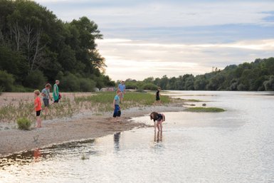 Eine Gruppe von Kindern spielt am Ufer eines ruhigen Flusses, während die Sonne hinter den Bäumen untergeht.