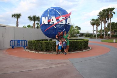 Eine Familie posiert fröhlich vor dem NASA-Symbol, umgeben von Palmen und einem gepflegten Garten.
