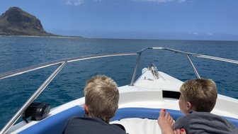 Zwei Jungen sitzen auf einem Boot und schauen auf das ruhige Wasser, während ein Berg im Hintergrund sichtbar ist.