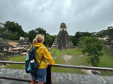 Eine Frau in gelber Jacke steht an einem Geländer und blickt auf die beeindruckenden Ruinen von Tikal unter einem bewölkten Himmel.