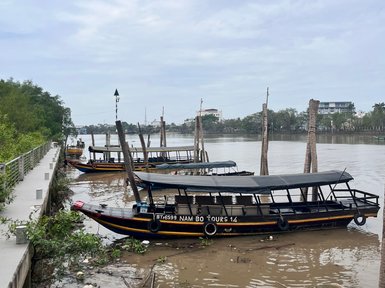 Drei traditionelle Boote liegen am Ufer des Mekong, umgeben von einer ruhigen Wasserlandschaft und sanften Wolken am Himmel.