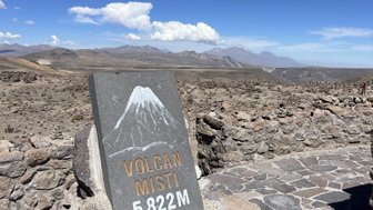Ein markanter Stein mit der Aufschrift 'VOLCAN MISTI 5822M' steht vor einer weiten, kargen Landschaft unter einem strahlend blauen Himmel.