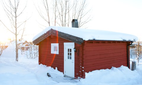Eine rote Holzhütte steht in einer verschneiten Landschaft, umgeben von kahlen Bäumen und sanften Hügeln.