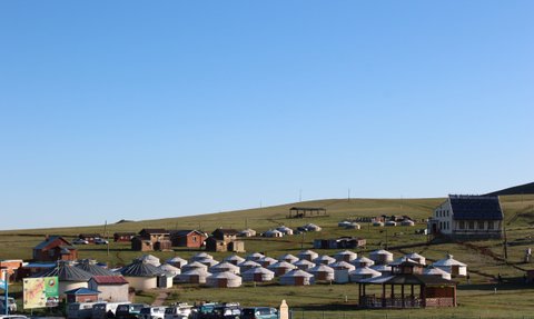 Eine weite Landschaft mit traditionellen Jurten und modernen Gebäuden, umgeben von sanften Hügeln unter klarem Himmel.