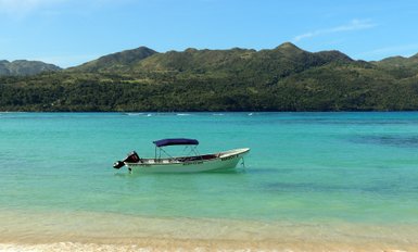 Ein kleines Boot mit einem blauen Sonnendach schwimmt ruhig im klaren, türkisfarbenen Wasser vor einer grünen Berglandschaft.