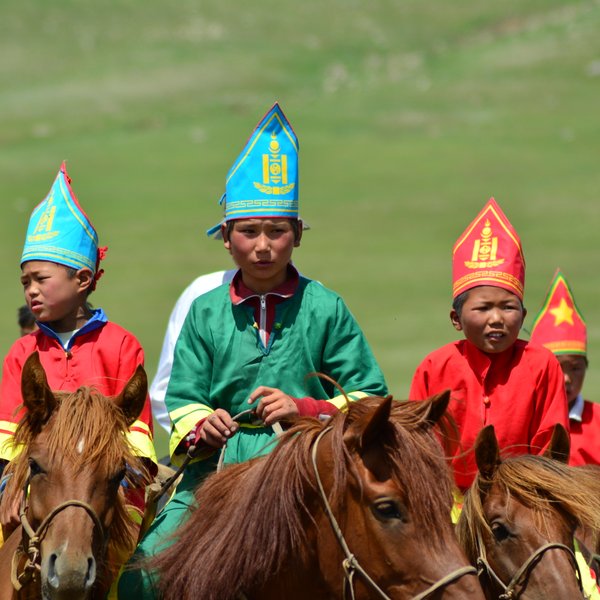 Eine Gruppe von Kindern in traditionellen bunten Gewändern reitet auf Pferden über eine grüne Wiese, voller Freude und Energie.