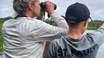 Eine Frau mit blonden Haaren beobachtet mit einem Fernglas die Umgebung, während ein Junge auf das Wasser zeigt.