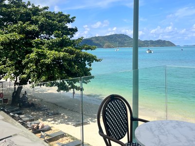 Ein Blick auf den Strand von einem Balkon mit einem Tisch und einem Stuhl, umgeben von Palmen und klarem Wasser.