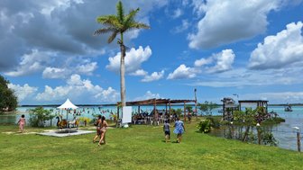 Ein sonniger Tag am Bacalar-See, mit Menschen, die am Ufer spazieren und im Wasser spielen, umgeben von üppigem Grün.