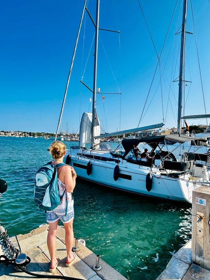 Eine Frau mit einem Rucksack steht am Hafen und beobachtet ein Boot, das im klaren Wasser anlegt.