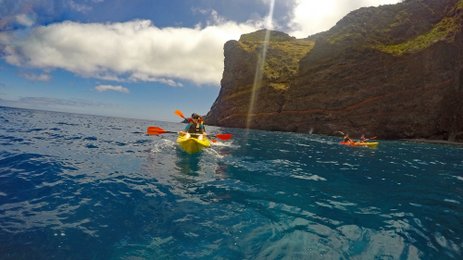 Zwei Kajakfahrer paddeln in klarem Wasser vor einer beeindruckenden Felsenküste unter einem strahlend blauen Himmel.