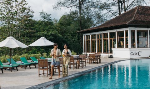 Ein Paar schlendert am Rand eines glitzernden Pools, umgeben von grünen Liegen und Sonnenschirmen, unter einem blauen Himmel.