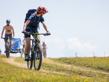 Eine Radfahrerin mit rotem Helm und Rucksack fährt auf einem schmalen Weg durch eine blühende Wiese.