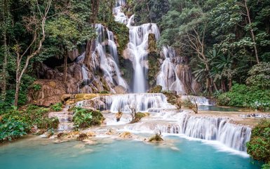 Ein majestätischer Wasserfall stürzt über mehrere Stufen in ein türkisfarbenes Becken, umgeben von üppigem Grün.