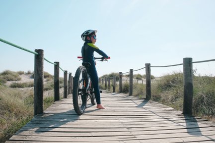 Ein Junge in einem blauen Neoprenanzug steht auf einem Holzsteg und schaut in die Ferne, während er sein Mountainbike hält.