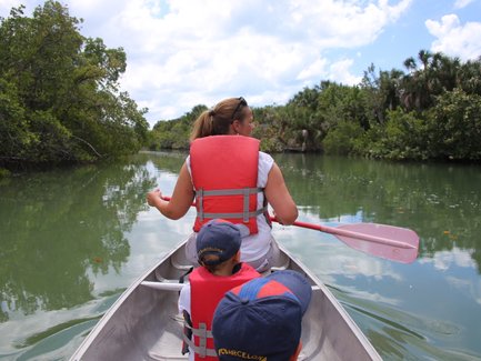 Eine Frau paddelt in einem Kanu durch ruhiges Wasser, umgeben von üppigem Grün und einem klaren Himmel.