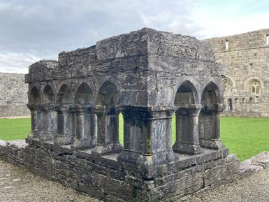 Ein historisches Steingebäude mit Bögen und Säulen, umgeben von grünem Gras und alten Mauern im Hintergrund.
