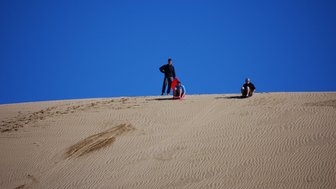 Drei Personen stehen und sitzen auf einer großen Sanddüne unter einem strahlend blauen Himmel, bereit für ein Abenteuer.