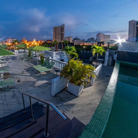 Eine Dachterrasse mit Liegen und Sonnenschirmen, umgeben von Pflanzen, bietet einen Blick auf die Stadt bei Dämmerung.
