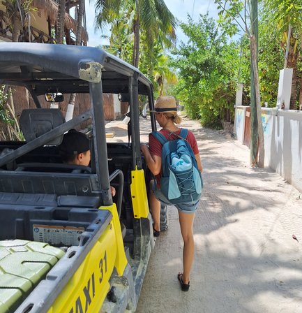 Eine Frau mit einem Rucksack und einem Hut nähert sich einem gelben Taxi auf einer sonnigen Straße, umgeben von Palmen.