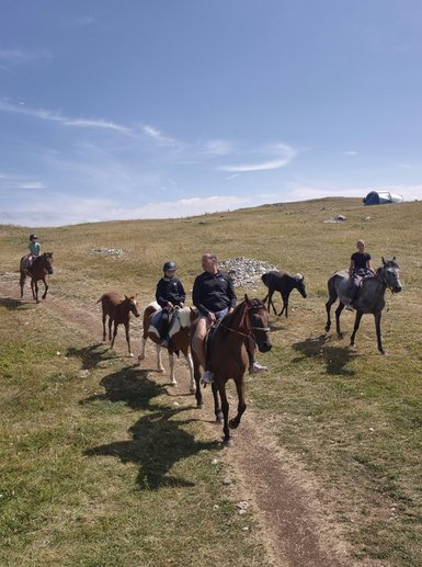 Familie reitet auf Pferden über eine malerische Wiese, umgeben von sanften Hügeln und blauem Himmel.