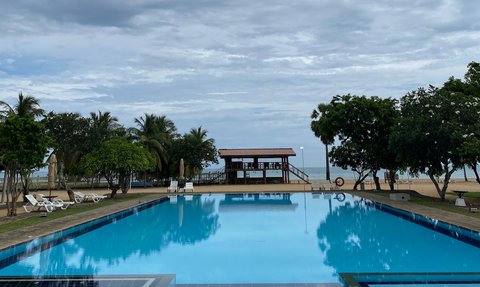 Ein weitläufiger Pool spiegelt den bewölkten Himmel wider, umgeben von Palmen und einer einladenden Liegewiese.