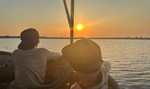 Zwei Personen sitzen auf einer Terrasse und beobachten den Sonnenuntergang über dem Wasser, umgeben von einer friedlichen Landschaft.