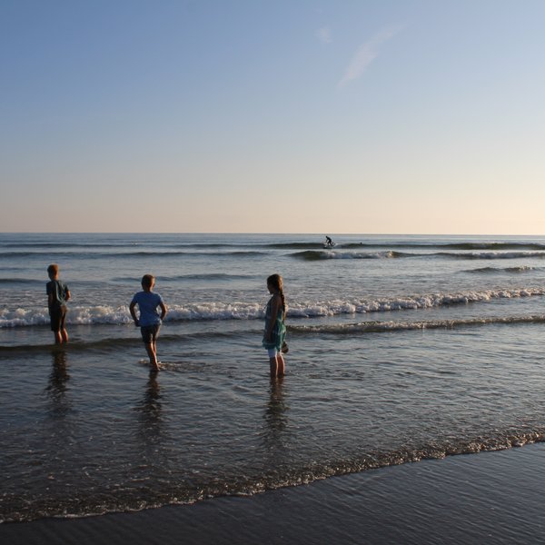 Drei Kinder stehen im flachen Wasser, während sanfte Wellen an den Strand rollen und die Sonne am Horizont scheint.