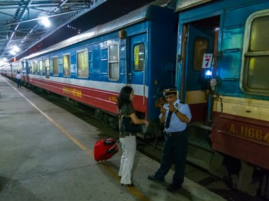 Eine Frau mit einem Koffer spricht mit einem Zugbegleiter an einem Bahnsteig in der Nacht.