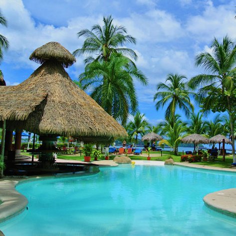 Ein einladender Poolbereich mit Palmen und einem Strohdach, umgeben von üppigem Grün und blauen Himmel.