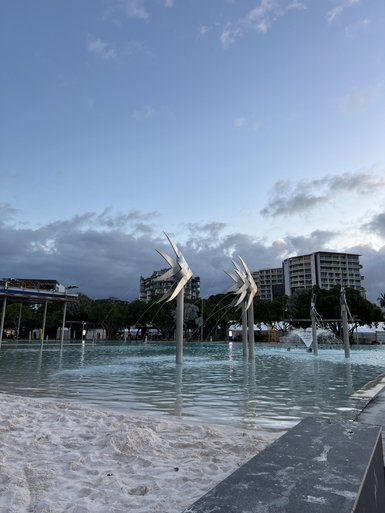 Ein ruhiger Lagunenbereich mit Kunstinstallationen und feinem Sand, umgeben von Bäumen und Wolken am Abend.