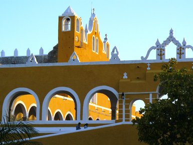 Die leuchtend gelbe Fassade eines historischen Gebäudes mit weißen Verzierungen und einem Uhrenturm im Hintergrund.