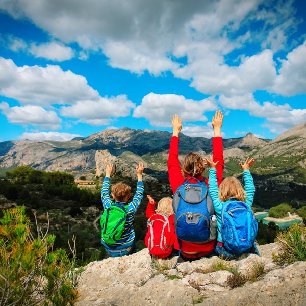 Eine Gruppe von vier Personen, darunter Kinder, genießt die Aussicht auf eine beeindruckende Berglandschaft und hebt die Hände.
