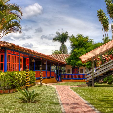 Ein farbenfrohes Haus mit roten und blauen Fenstern, umgeben von üppigem Grün und Palmen, unter einem klaren Himmel.