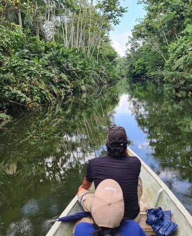 Zwei Personen sitzen in einem Boot auf einem ruhigen Fluss, umgeben von üppigem, grünem Dschungel und spiegelndem Wasser.
