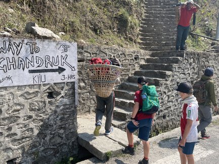 Eine Gruppe von Kindern steht an einer steinernen Treppe, während ein Erwachsener mit einem Korb auf dem Rücken vorbeigeht.