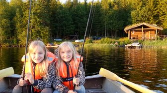 Zwei fröhliche Mädchen in orangefarbenen Schwimmwesten sitzen im Boot und halten Angelruten in der Hand.