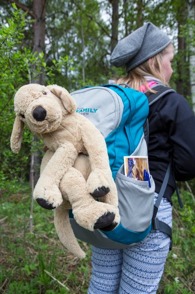 Ein Mädchen mit einem Rucksack, auf dem ein plüschiger Hund sitzt, steht in einer grünen Waldlandschaft.