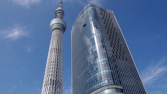 Der Tokyo Skytree erhebt sich majestätisch über die moderne Architektur eines angrenzenden Wolkenkratzers unter blauem Himmel.