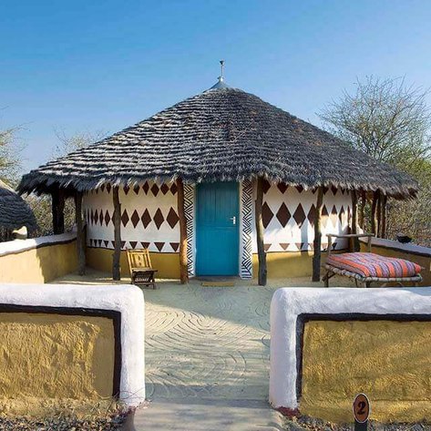 Ein traditionelles Rundhaus mit einem strohgedeckten Dach und einer blauen Tür, umgeben von einer sandigen Terrasse.