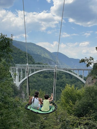 Zwei Kinder sitzen zusammen auf einer Schaukel und genießen den Blick auf die beeindruckende Landschaft und Brücke.