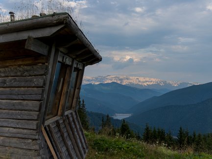 Ein schief stehendes Holzgebäude mit großen Fenstern bietet einen atemberaubenden Blick auf die bewaldeten Berge im Hintergrund.