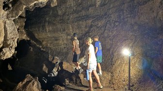Drei Personen erkunden eine Höhle, umgeben von rauen Felswänden und schwachem Licht, während sie auf unebenem Boden stehen.