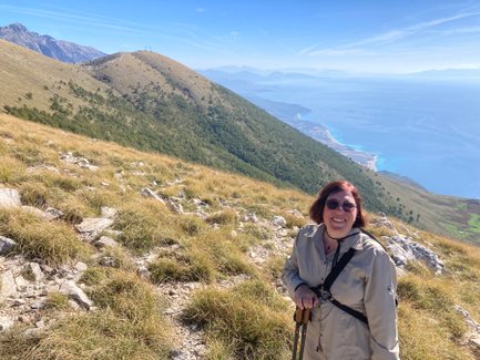 Eine fröhliche Frau mit Sonnenbrille steht auf einem Berg, umgeben von grünen Hügeln und dem blauen Meer im Hintergrund.