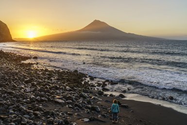 Eine Person steht am Strand und betrachtet den Sonnenuntergang über dem Meer, während der Vulkan im Hintergrund sichtbar ist.
