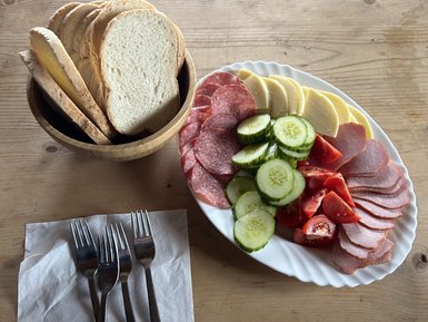 Ein rustikaler Holztisch mit einer Schüssel Brot und einem Teller mit Wurst, Käse, Gurken und Tomaten.