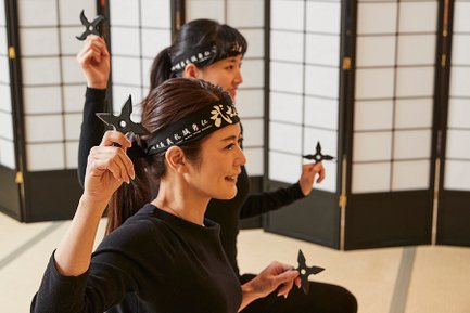 Zwei Frauen in schwarzer Kleidung halten Ninja-Sterne und lächeln, während sie in einem traditionellen Raum stehen.